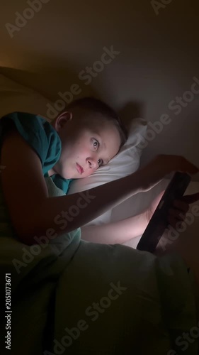 Vertical. A child holds a smartphone under the blanket's light, scrolling through content. The boy uses gadgets while getting ready for bed, his face illuminated by the blue light of the smartphone sc