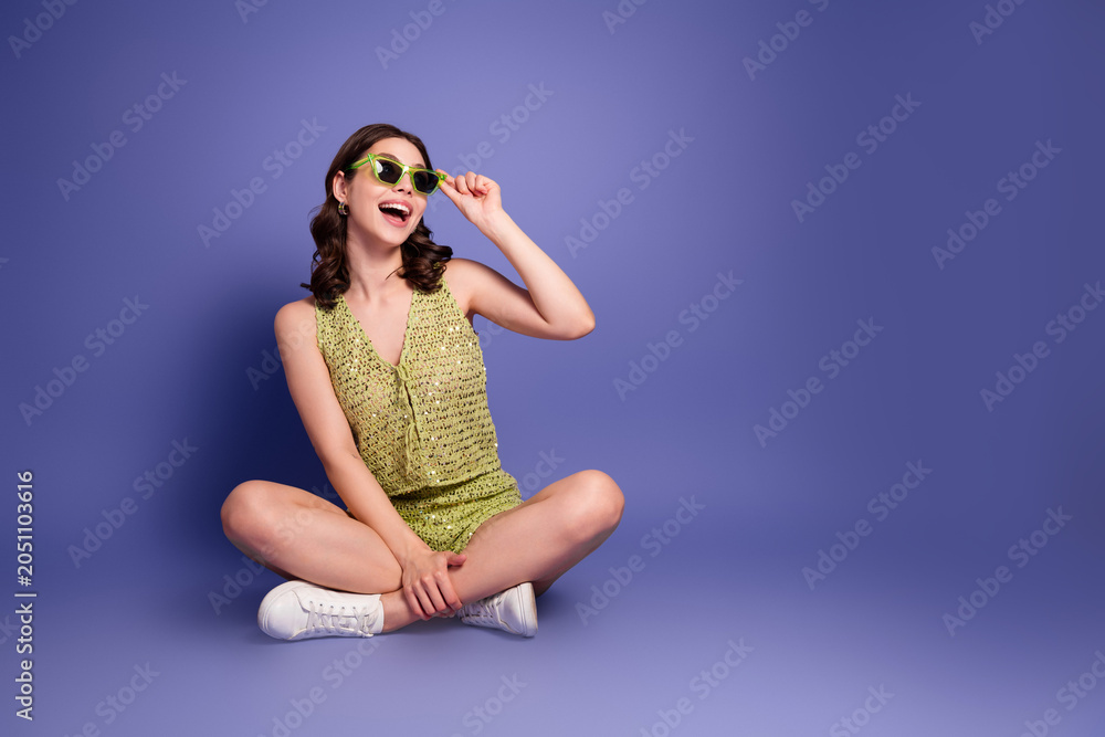 Fototapeta premium Happy young woman sitting cross-legged on purple background wearing green summer outfit and sunglasses