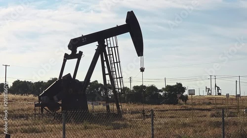 Oil pumpjack silhouette works in a dry field with fences power lines and industrial equipment, Editorial video frame shows petroleum extraction machinery marked with visible numbers in rural landscape