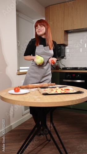 Happy young woman makes pizza at home. Cheerful red-haired girl in apron holds fresh lettuce and tomato in kitchen, posing for photo. Healthy cooking, culinary hobbies. Cooking with pleasure