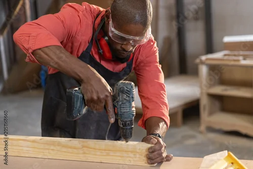Fototapeta Cordless drill driving screw into wooden plank on workbench in workshop with scattered wood panels