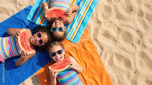 Three girls lay on colorful towels on a sandy beach. They wear sunglasses and hold large slices of watermelon while smiling at the camera on a sunny summer day