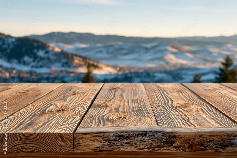 Naklejka premium Rustic wooden table surface with a soft focus background of snow-covered mountains at sunset.