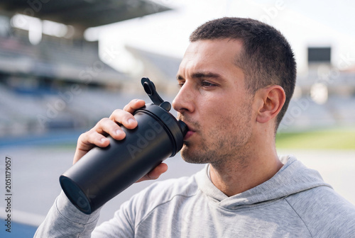 A fit young man in a light grey hoodie drinking protein coffee from a matte black shaker on a blurred stadium background, sunny day, fitness nutrition and healthy lifestyle concept