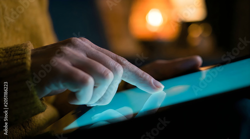 Close up of person's hand with pointer finger touching glowing screen of digital tablet in dimly lit room.