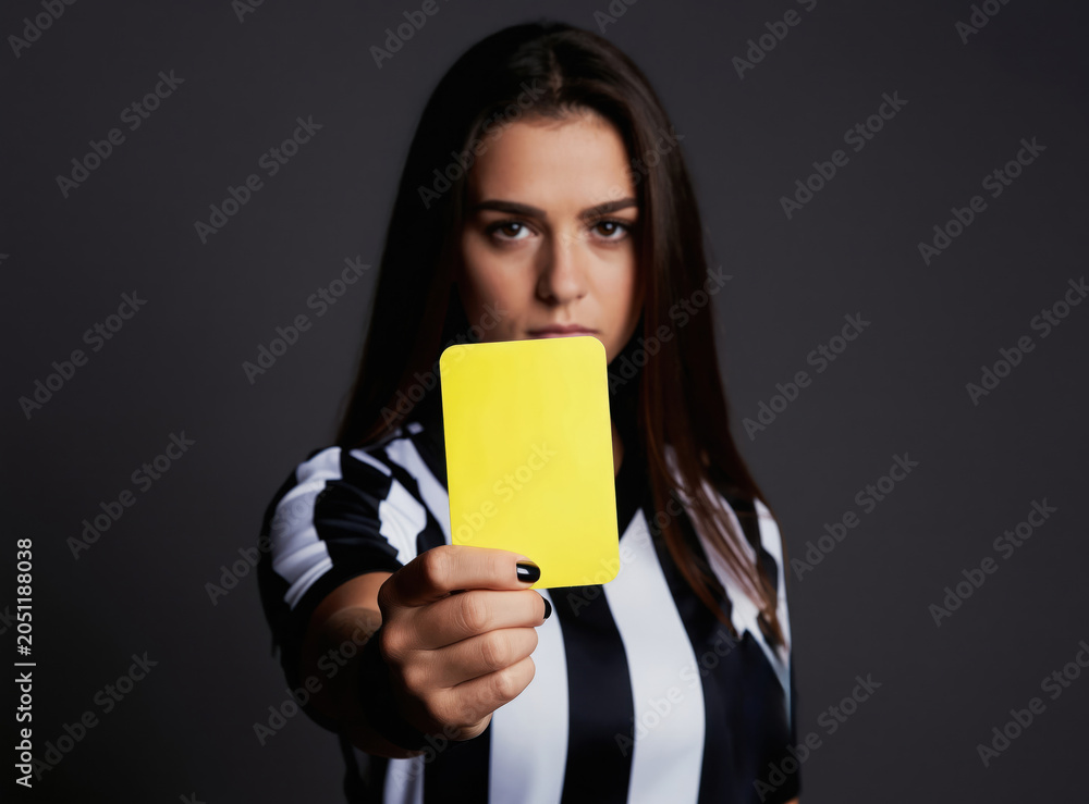 Naklejka premium Young woman soccer referee in striped uniform shows yellow card. Serious female ref holds warning card for player foul in studio. Sport game official gives penalty.