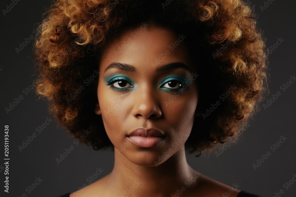 Fototapeta premium Young Black woman with afro hairstyle wears bright teal eye makeup. She looks forward with a neutral expression. Her curly hair has blonde highlights. Studio portrait.