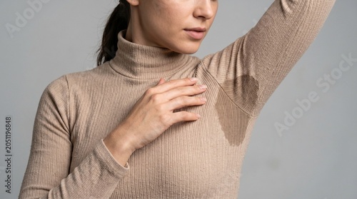 A woman wears a tight beige turtleneck sweater showing a noticeable dark sweat stain under her arm in bright lighting