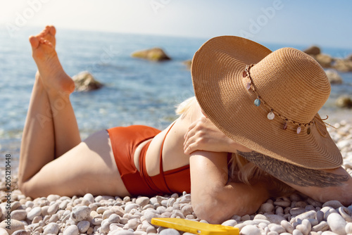 Fotografija Woman sunbathing on pebbled shore wearing straw hat, lying back in swimsuit and