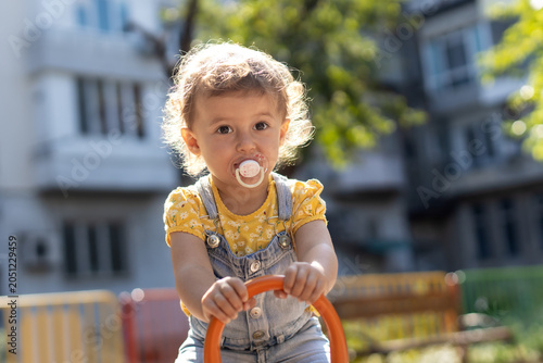 Adorable toddler girl with a pacifier and curly hair playing at an outdoor playground on a sunny day.