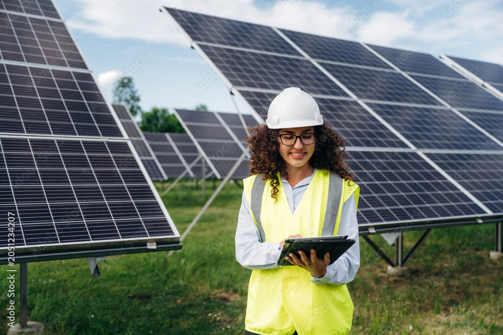 Obraz premium Female engineer wearing safety vest and helmet using tablet in solar farm with solar panels and green grass under blue sky on sunny day