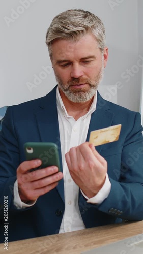 Middle aged businessman programmer software developer working making online purchase payment shopping with credit bank card smartphone at modern office desk with laptop. Mature freelancer business man