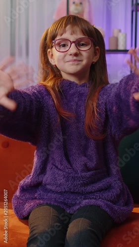 Girl smiling friendly at camera and waving hands gesturing hello, hi, greeting or goodbye, welcoming with hospitable expression. Child kid at home in night evening living play room on couch. Vertical