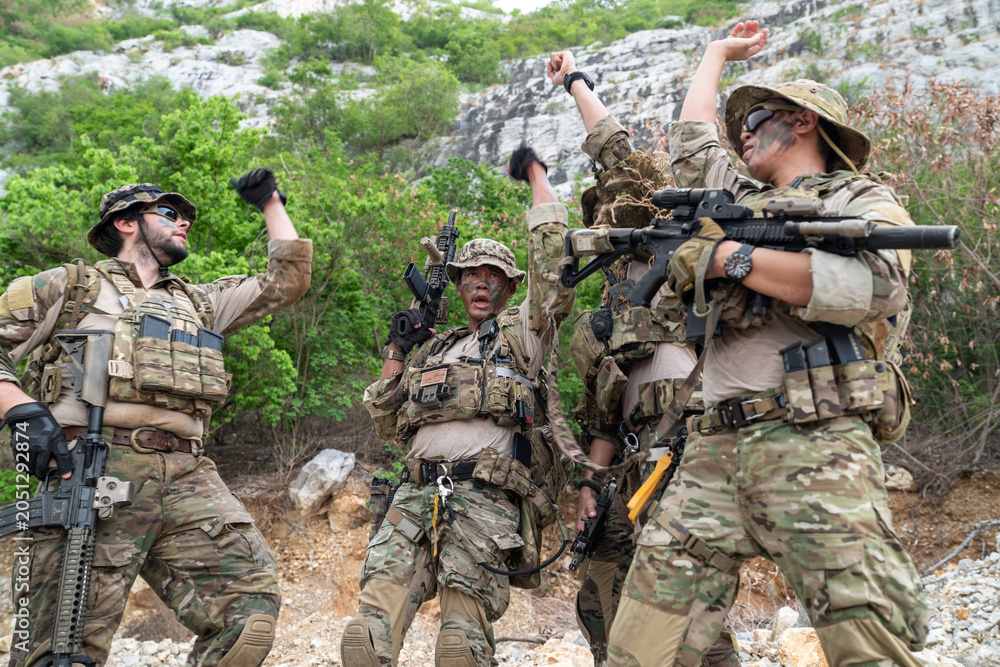 Fototapeta premium Group of four soldiers in camouflage uniform stacking hands during military tactical briefing outdoor, special forces operators showing team unity near rock cliff
