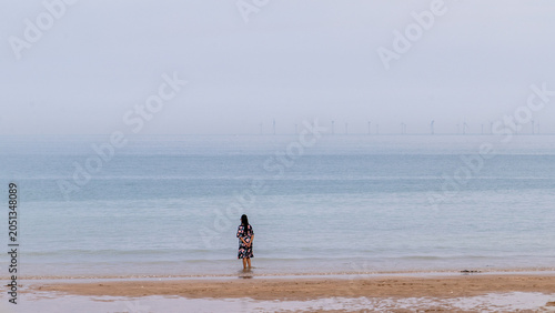 Woman standing on calm misty beach.Calm beach scene with a lone woman at the shoreline and distant paddle boarders on a misty sea under a soft cloudy sky.