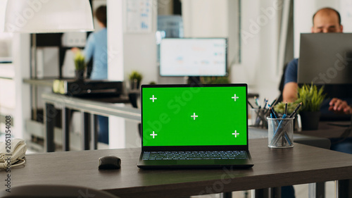 Fotografie Green screen laptop in coworking space placed on empty office desk, open floor plan business workplace with isolated display, with portable computer running blank chroma key layout on software