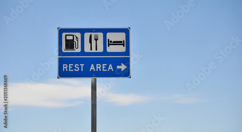 Blue highway rest area road sign with gas, food, and lodging icons against a clear blue sky