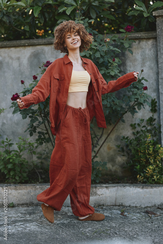 Photos A young woman with curly hair twirls in an orange pantsuit and cream top in a su