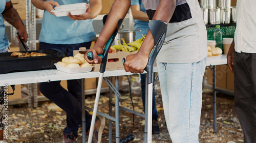 Fotografija African American woman volunteer assists and supports crippled man at outdoor food bank, with the hungry, poor and homeless people receive hunger relief assistance from multiethnic voluntary