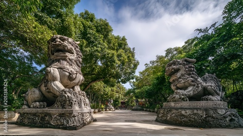 Two ancient stone lion statues guard the entrance to a serene pathway lined with lush green trees and dappled sunlight. The statues, weathered by time, emanate a sense of history and mystery.