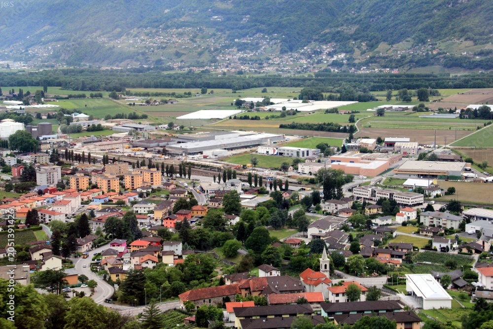 Fototapeta premium Aerial view of a vibrant town with red rooftops, church tower, railway yard, and green mountains blending urban life with natural landscapes.