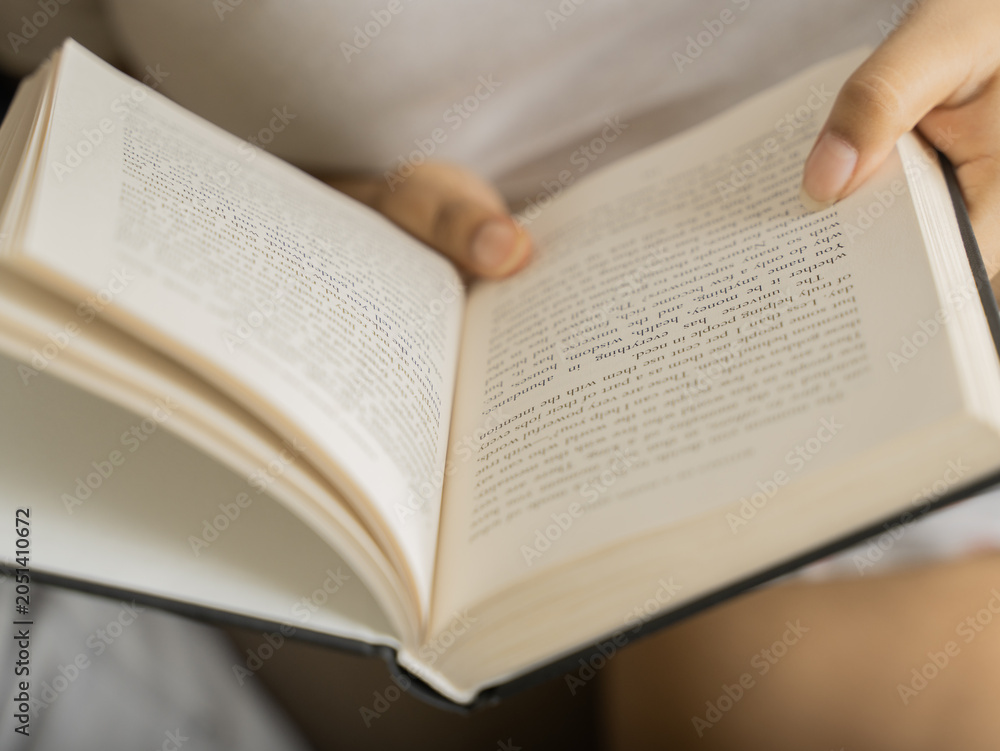 Fototapeta premium Close-up of a woman's hands holding an open book with clearly visible text pages, shot in warm cozy indoor lighting with a soft glowing table lamp in the background, evoking reading and knowledge. 