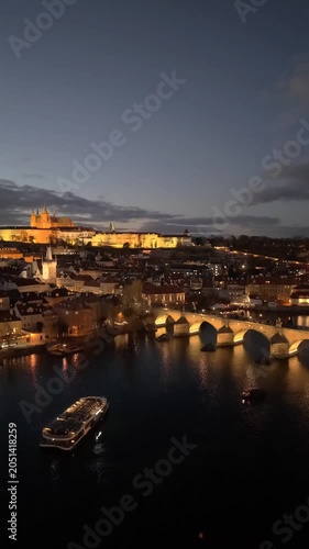 Aerial views capture the beauty of Prague as the city lights up at dusk. Iconic landmarks like Prague Castle and Charles Bridge glow against the darkening sky along the Vltava River.