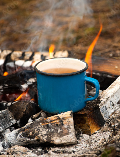 Blue enamel mug of hot beverage steaming over a campfire in the woods
