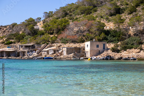 Ibiza, Spain, Crystal-clear turquoise water of Cala Vedella bay washing against a rocky shore with traditional fisherman huts in the background, showing the transparent Mediterranean sea.