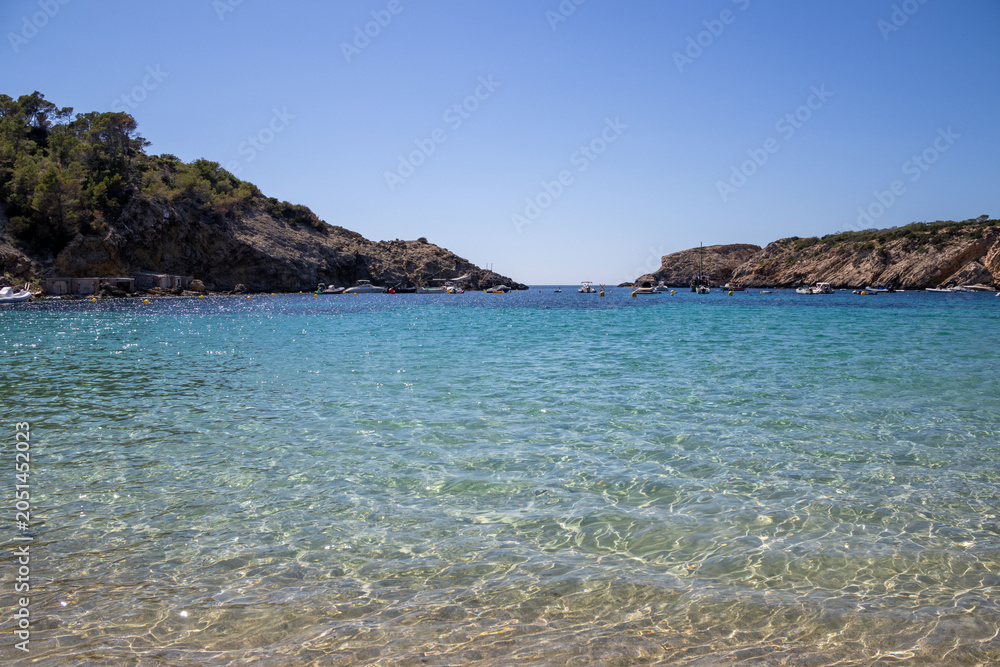 Obraz premium Ibiza, Spain. Crystal-clear turquoise water of Cala Vedella bay with small boats anchored near the rocky shore, showcasing the transparent sea and sandy bottom.