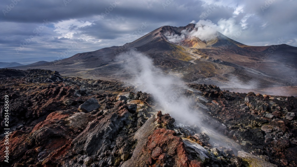 Fototapeta premium Active volcanic landscape with steaming fumaroles, rugged lava fields and a smoking crater beneath a dramatic cloudy sky