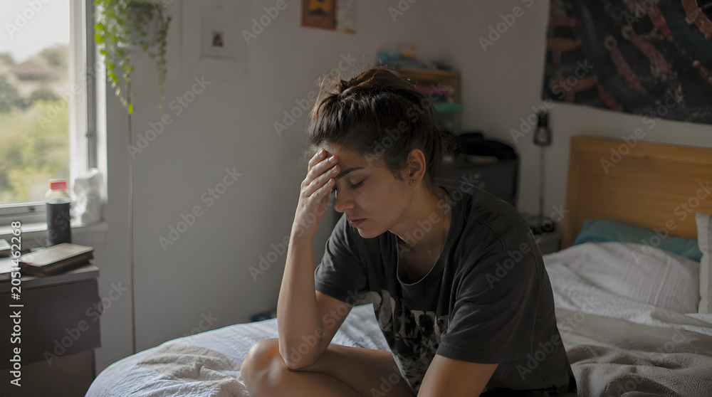Fototapeta premium A young woman sits on her bed with her hand on her forehead, appearing distressed or lost in thought. The natural light from the window illuminates the scene, creating a somber mood.