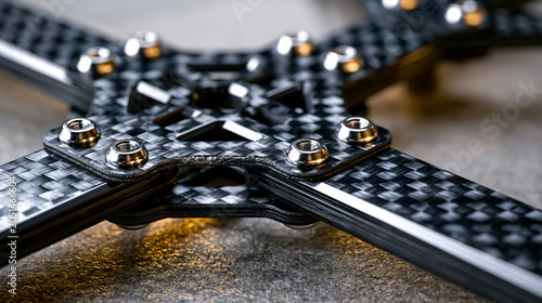 Quadcopter drone frame section in woven carbon fiber with motor mount arm junction, countersunk M3 titanium bolt pattern and ply edge delamination macro visible, frame standoff hardware detail, ligh