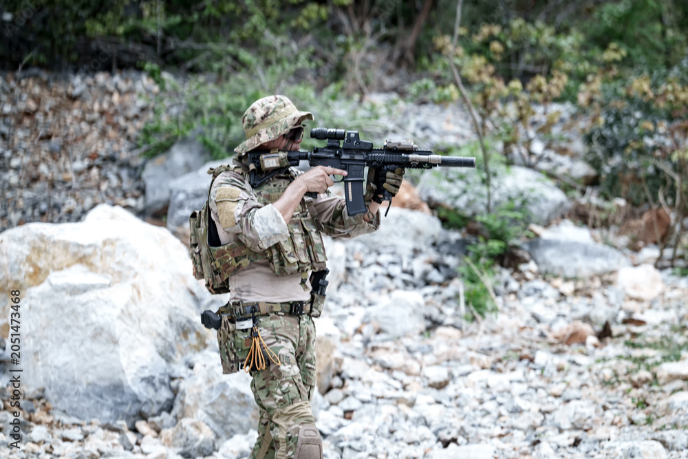 Fototapeta premium Soldier in camouflage gear aims rifle in rocky, wooded terrain