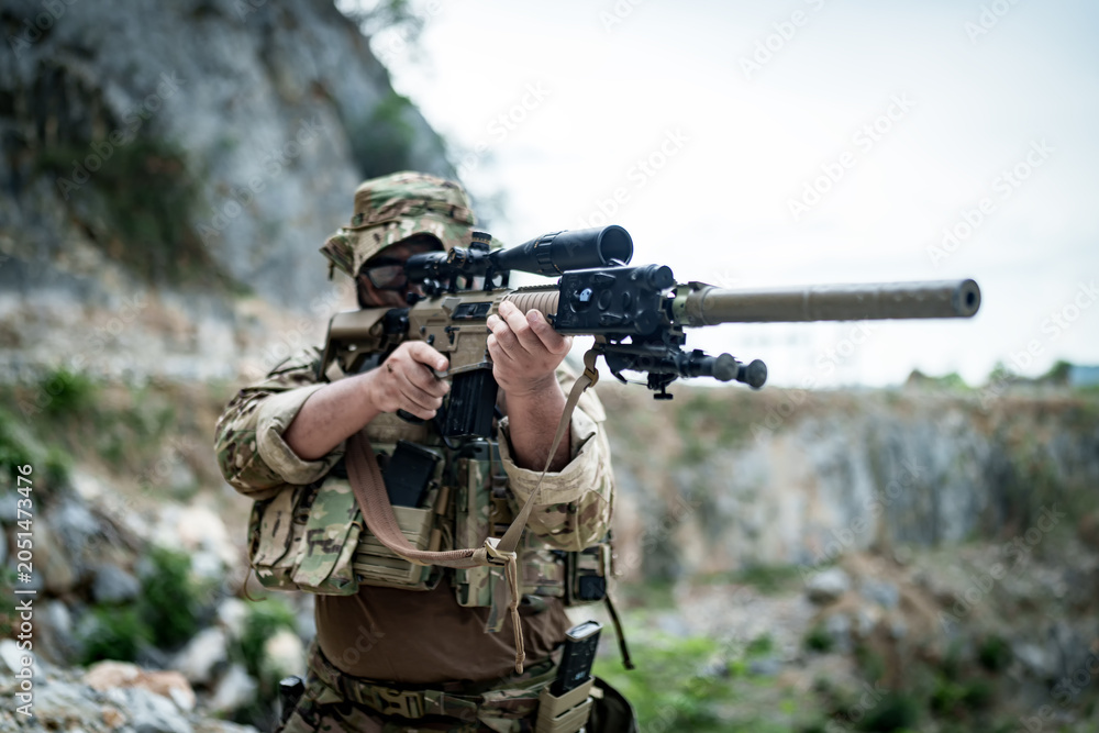 Fototapeta premium Soldier aiming a sniper rifle with scope and suppressor in a rocky outdoor environment