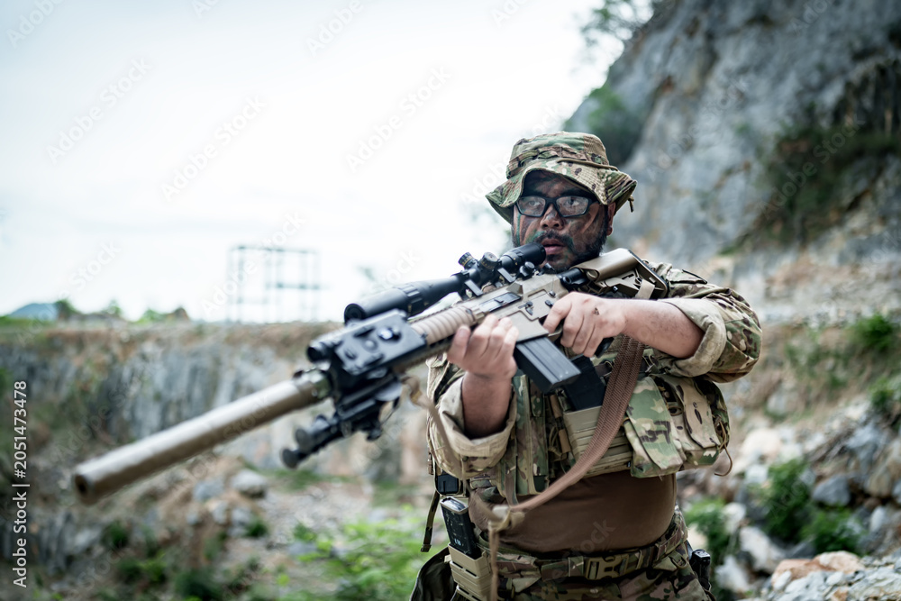 Fototapeta premium Soldier in camouflage gear aims a sniper rifle with a scope in a rocky outdoor setting
