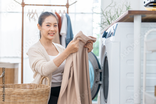 Asian woman, laundry, washing machine, clothing, home, basket smiling while loading fabric into front load appliance in bright utility room interior