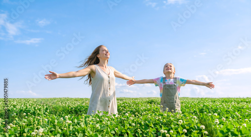Happy mother and daughter with arms outstretched in a green clover field. Joyful family embracing nature under a blue sky