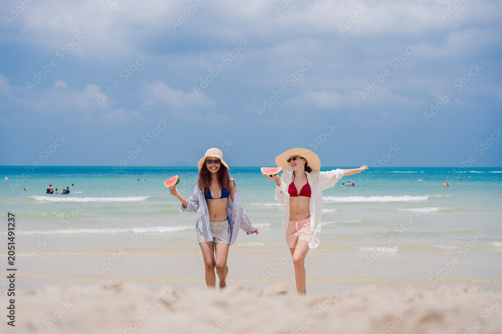 Fototapeta premium Summer beach friendship concept. Two happy women in swimwear enjoying fresh watermelon on tropical beach, celebrating vacation freedom and sunny holiday moments. summer, women, vacation, friendship