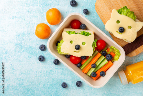 Kids school lunch box with funny fox sandwich with cheese, and fresh vegetables, carrot, tomatoes, celery, blueberries and apricots, orange juice. Back to school background. Top view, blue background.