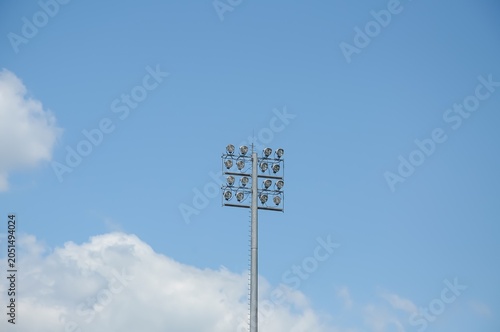 Tall stadium light tower with multiple floodlights against a clear blue sky and white clouds, outdoor sports infrastructure