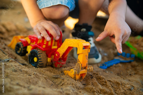 Under the evening sun, a child plays in a sandbox with a toy plastic red excavator. Close-up photo without a face, focus on an excavator toy car.