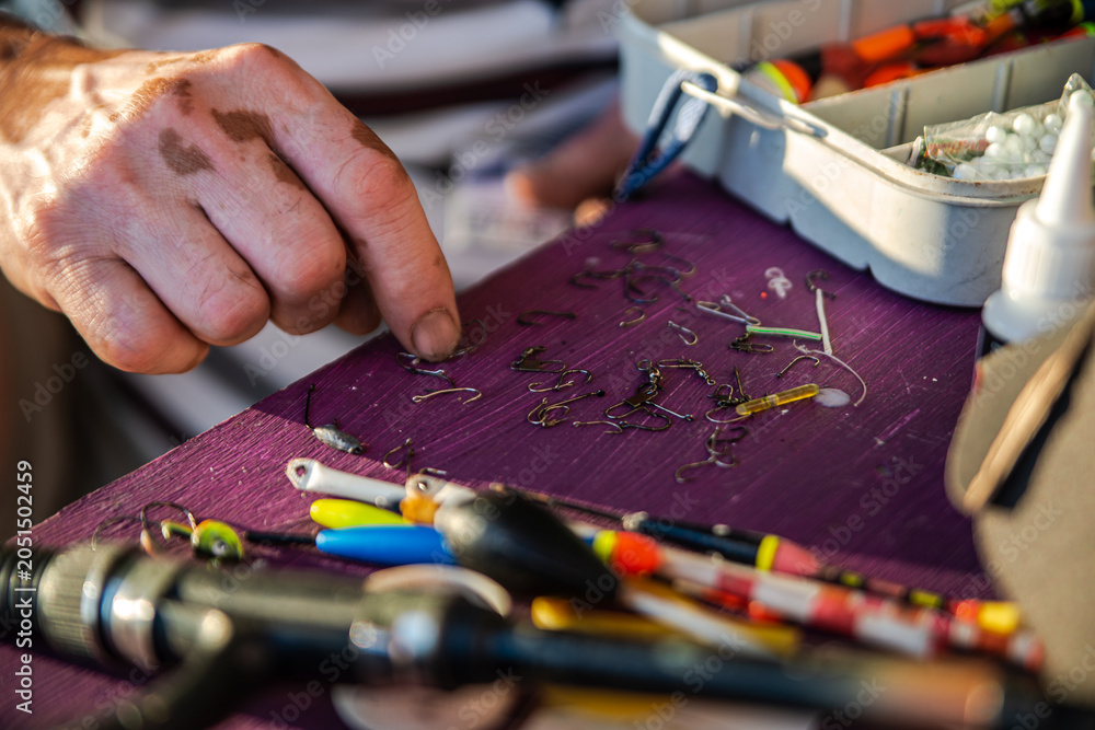 Fototapeta premium A person lays out fishing gear, mainly hooks, on a purple surface. Fishing floats, sinkers, a spinning rod, a fishing box and a tube of glue are visible.