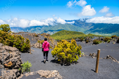 Hiker in a bright pink jacket walks along a black volcanic ash trail looking out over lush green valleys near Tajogaite volcano, La Palma, Canary Islands, Spain