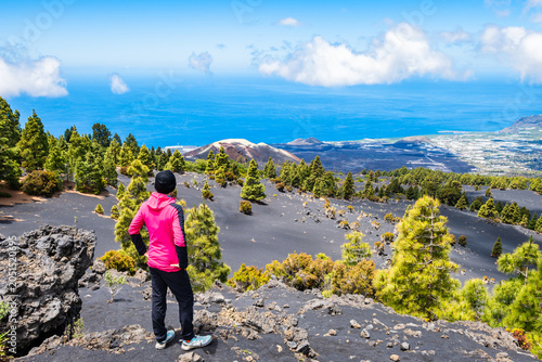 Hiker in a pink jacket stands on a volcanic trail looking down at a crater, green pines, and the blue sea near Tajogaite volcano, La Palma, Canary Islands, Spain