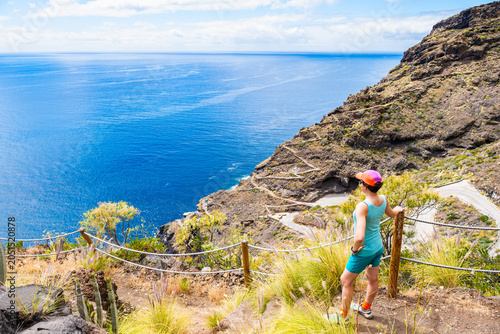 Female hiker stands at a cliffside overlook with rope railings, viewing a winding coastal road and blue ocean at Poris de la Candelaria, La Palma, Canary Islands, Spain