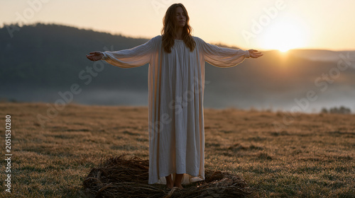 Woman standing barefoot on dew-covered grass at sunrise during a spring equinox ritual, arms open wide, mist still clinging to a distant treeline, a handmade wreath of branches