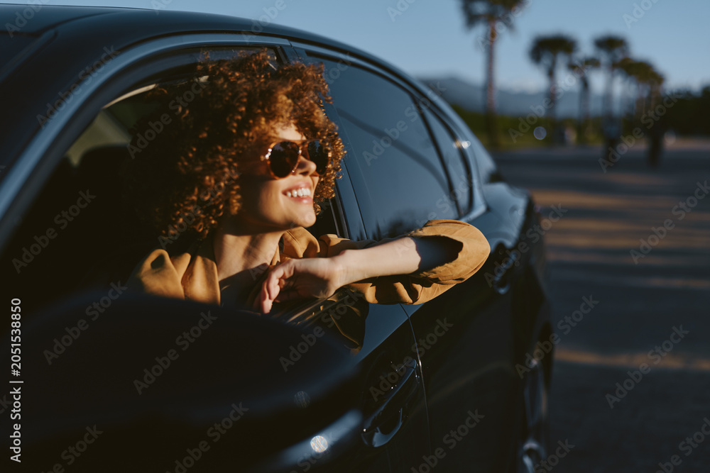 Fototapeta premium Smiling woman with curly hair leaning from a car window, enjoying a sunny drive. Retro film color mood enhances a carefree lifestyle and warm, optimistic vibe. Perfect for lifestyle scenes.