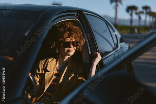 Fényképezés A stylish woman with curly hair in sunglasses sits in a vintage car, sunlit street ahead, embodying carefree road trip vibes and a retro lifestyle moment