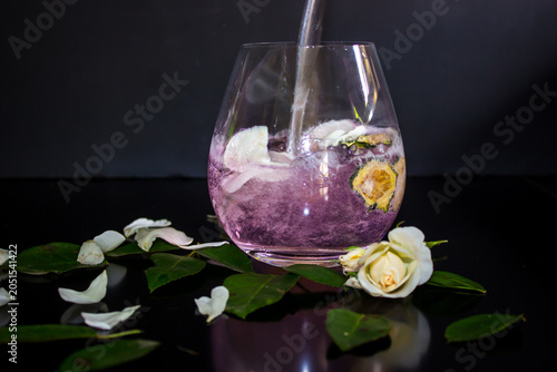 A rose flavored gin and tonic being poured in a glass with dried cucumber and rose petals.